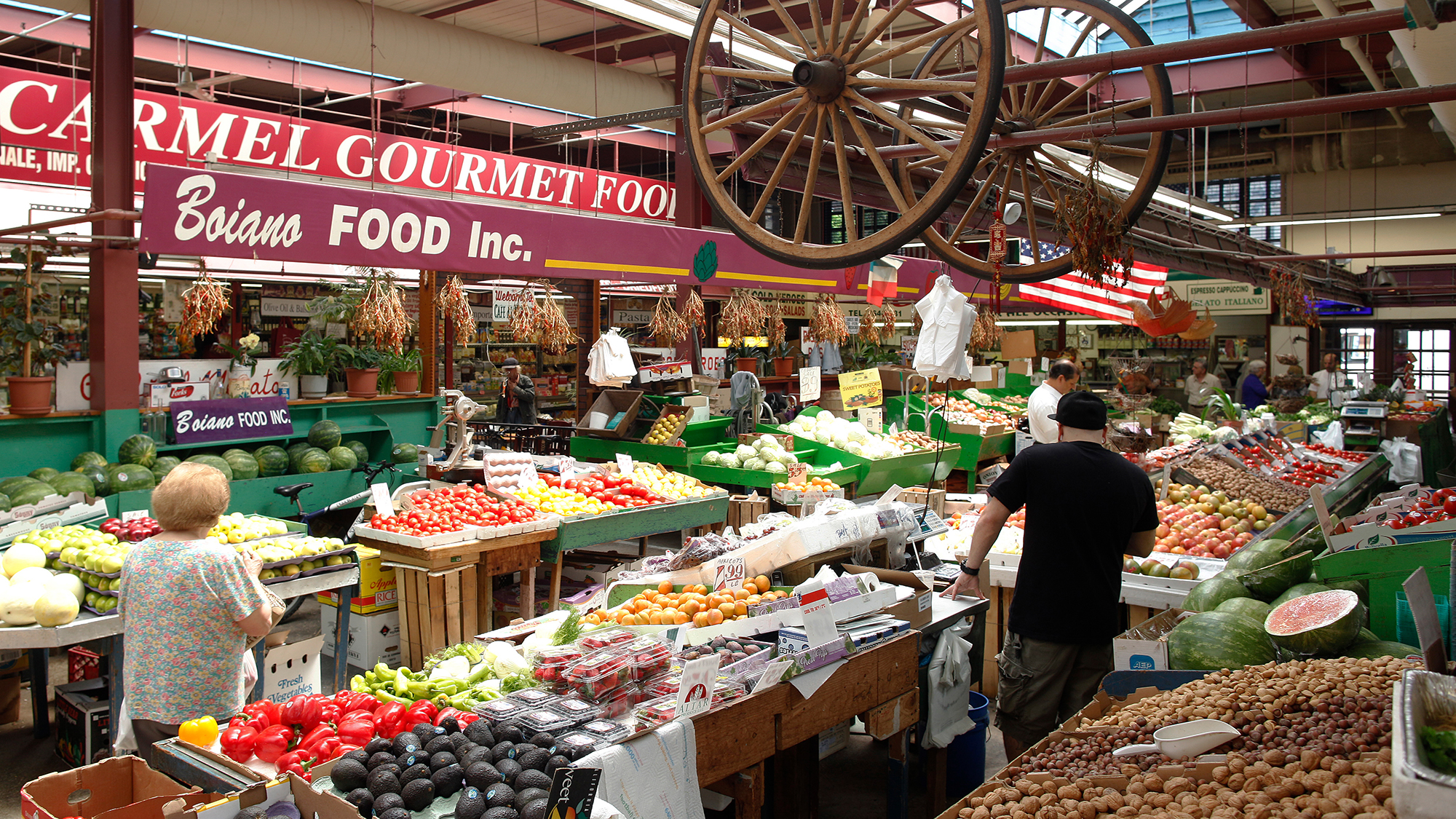 A busy indoor shot of Arthur Avenue Retail Market with various fresh fruits and vegetables displayed on tables. Shoppers browse the produce, and colorful signs hang above the stalls. A large wooden wagon wheel is suspended from the ceiling.