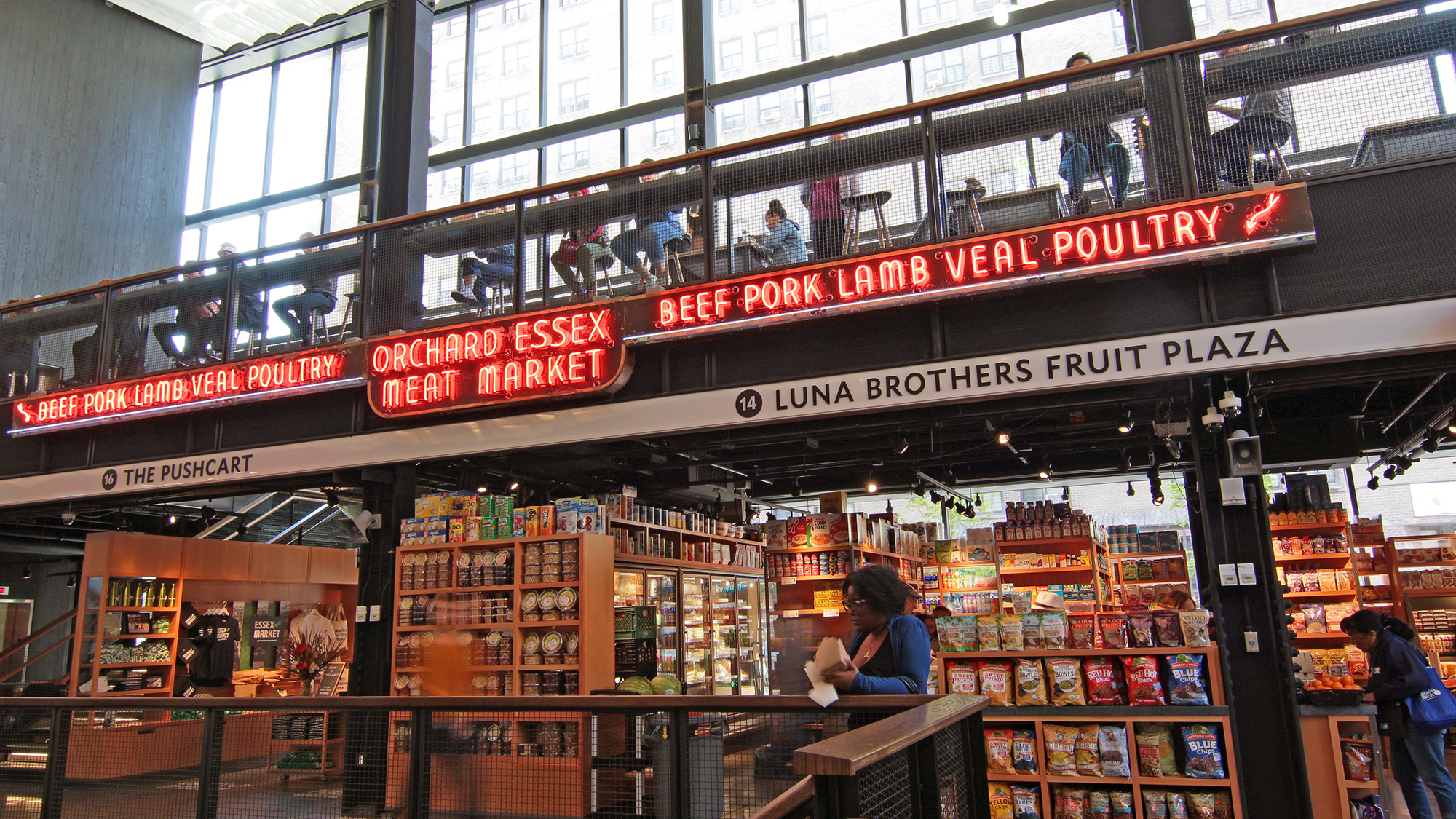 A spacious indoor market with shelves of groceries, neon signs reading “BEEF PORK LAMB VEAL POULTRY” and “ORCHARD ESSEX MEAT MARKET,” and people shopping or sitting on an upper level with large windows.