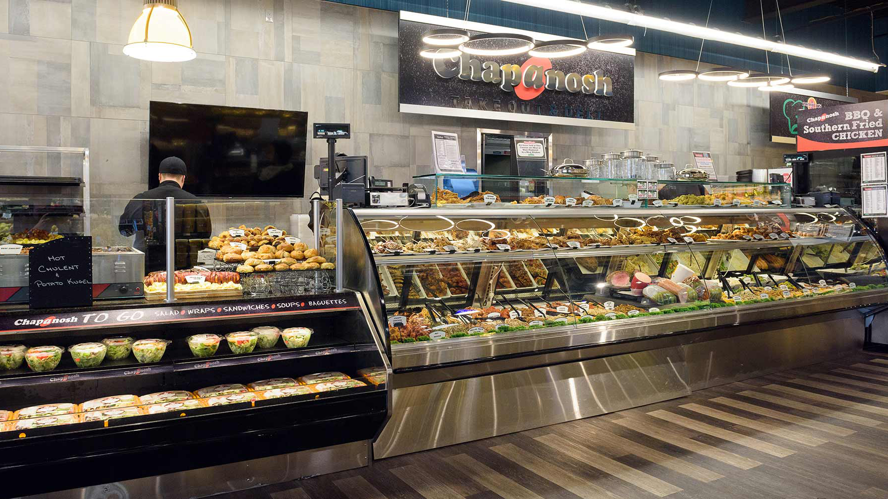 A deli counter displays salads, prepared meals, pastries, and hot foods. Behind the counter, a staff member assists. Signs read Chapnosh and BBQ & Southern Fried Chicken. The floor is wooden, and lighting is bright.