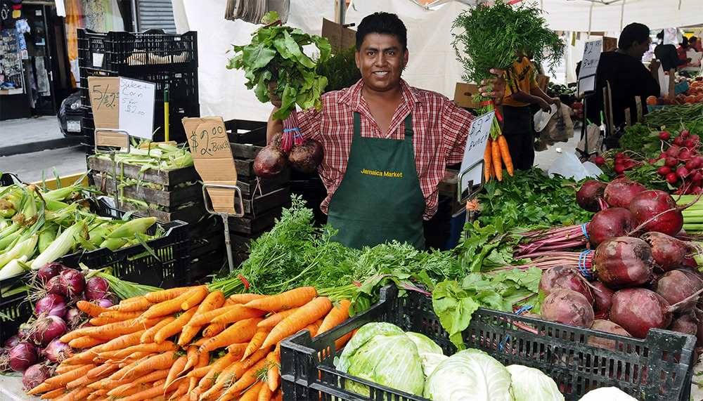 A smiling vendor in a green apron stands behind a market stall displaying carrots, beets, onions, lettuce, and other fresh vegetables, holding bunches of carrots and beets in each hand.