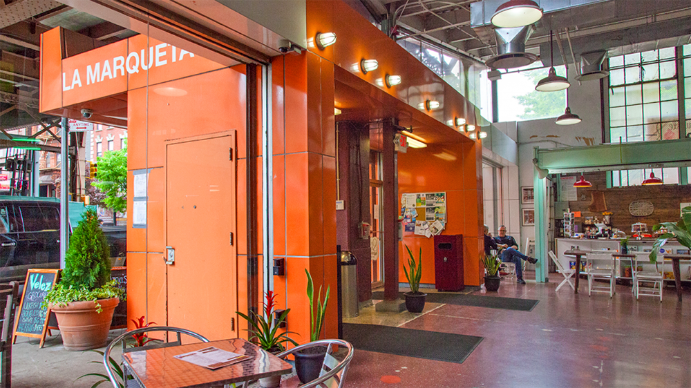 Indoor view of La Marqueta market with orange entrance, plants, bulletin board, small tables with chairs, and people sitting inside. Large windows let in natural light.