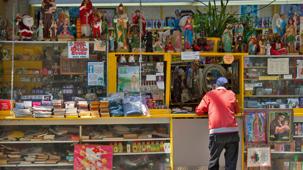 A person in a red jacket stands at a colorful outdoor shop counter filled with religious statues, candles, books, and various spiritual items displayed on shelves and hanging above.