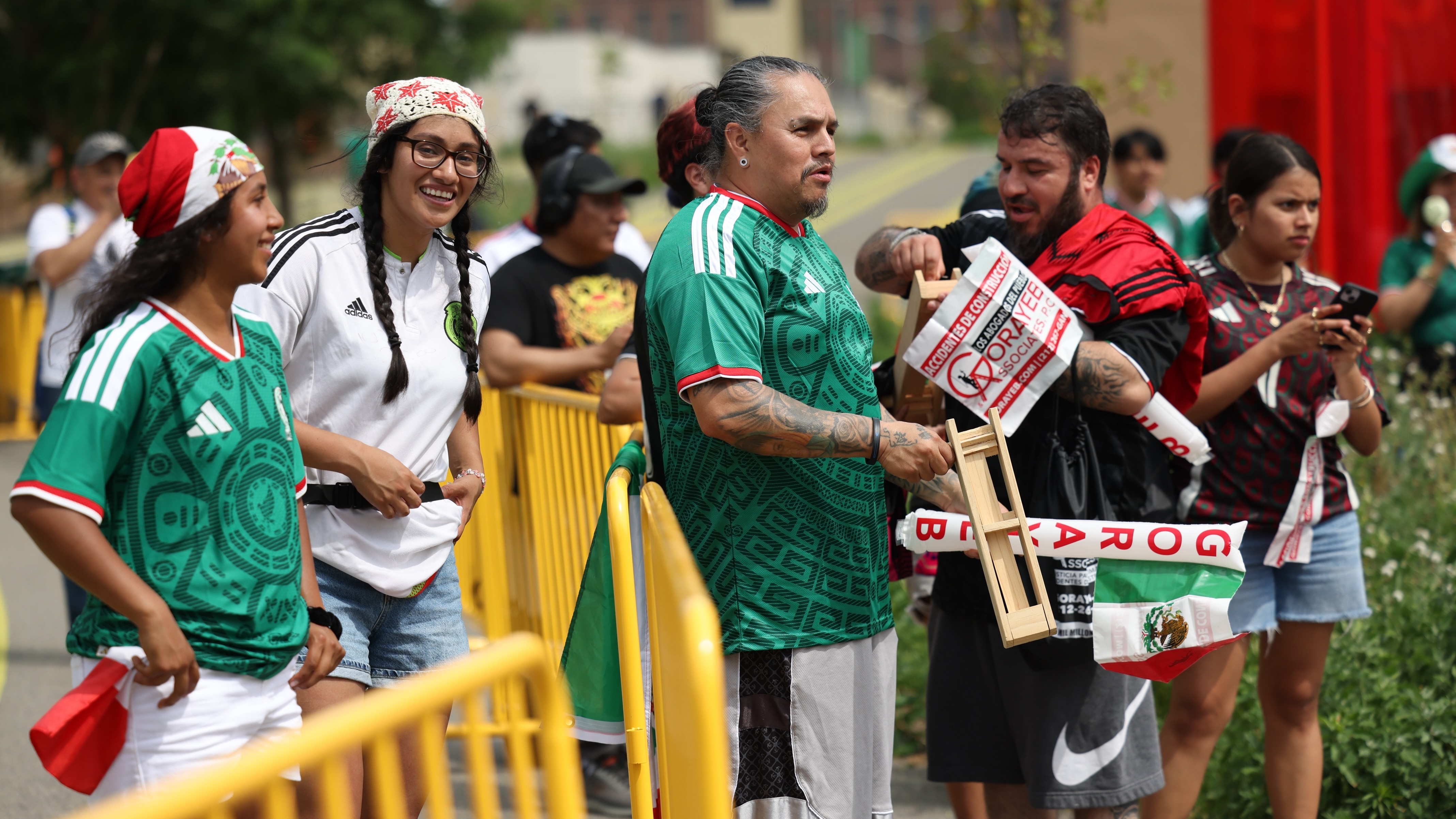 A group of soccer fans in green Mexico jerseys stand behind a yellow barrier, smiling and holding flags. One man holds a wooden noisemaker, and others wear festive hats and accessories. It is a festive outdoor setting.