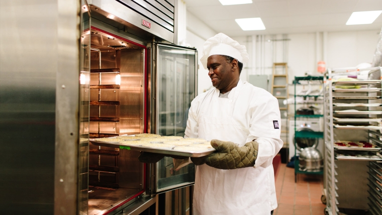 A chef from Hot Bread Kitchen wearing a white uniform and hat places a tray of food into a commercial oven. He is in a professional kitchen with industrial equipment and trays on racks in the background.