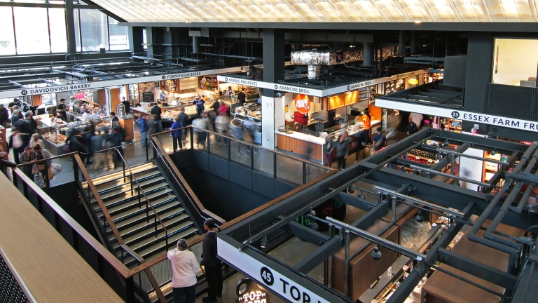 A bustling Essex Market with shops and food stalls. People explore different levels, accessed by stairs. The ceiling features a large, textured design. Glass windows let in natural light, and signage indicates various vendors, creating a vibrant atmosphere. Photo by Nina LoSchiavo.