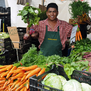 A smiling vendor in a green apron stands behind a market stall displaying carrots, beets, onions, lettuce, and other fresh vegetables, holding bunches of carrots and beets in each hand.
