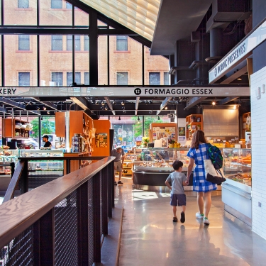 A woman and a child walk hand in hand through Essex Market, a bright and modern indoor market with bakery and cheese shop signs, glass displays of food, and large windows letting in natural light.