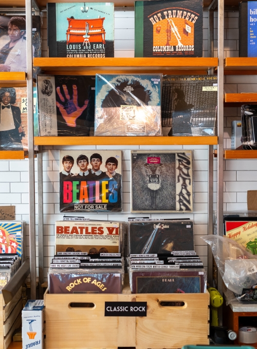 A display shelf holds an assortment of vinyl records. The top shelf features albums from bands and jazz artists. Below, a crate labeled Classic Rock contains various rock albums, including those by The Beatles, arranged in an organized manner. A display shelf holds an assortment of vinyl records. The top shelf features albums from bands and jazz artists. Below, a crate labeled Classic Rock contains various rock albums, including those by The Beatles, arranged in an organized manner.