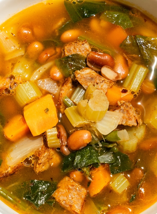 A close-up of a bowl of hearty vegetable soup with visible chunks of carrots, celery, leafy greens, beans, and pieces of meat in a rich broth. The soup appears colorful and appetizing.