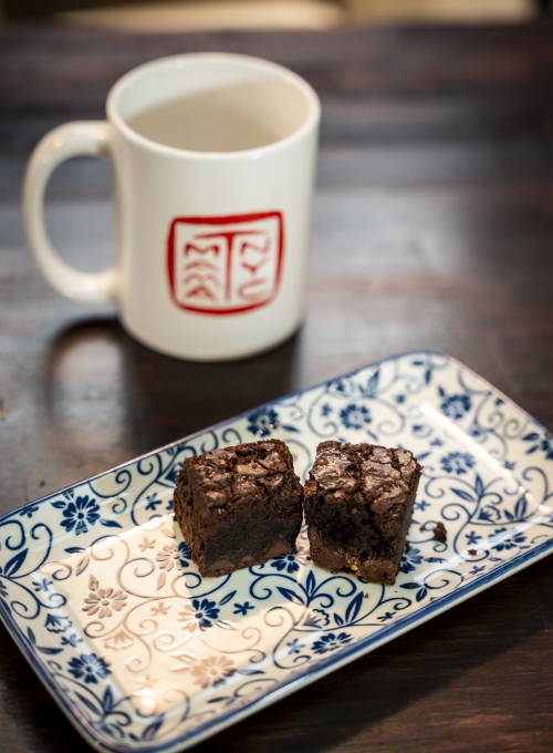 Two brownies on a blue and white floral-patterned plate, placed on a dark wooden table. A white mug with a red design is in the background. Two brownies on a blue and white floral-patterned plate, placed on a dark wooden table. A white mug with a red design is in the background.