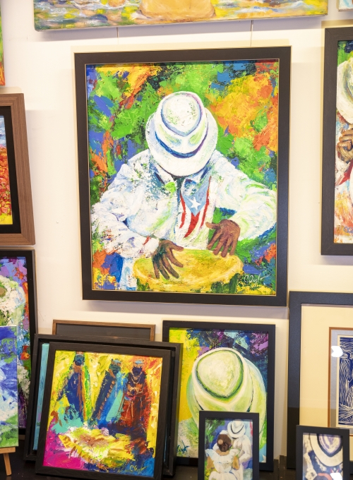 A vibrant display of colorful paintings, including a large framed painting of a figure in a white hat playing a drum. Other smaller paintings featuring abstract and colorful scenes are arranged around it. A vibrant display of colorful paintings, including a large framed painting of a figure in a white hat playing a drum. Other smaller paintings featuring abstract and colorful scenes are arranged around it.