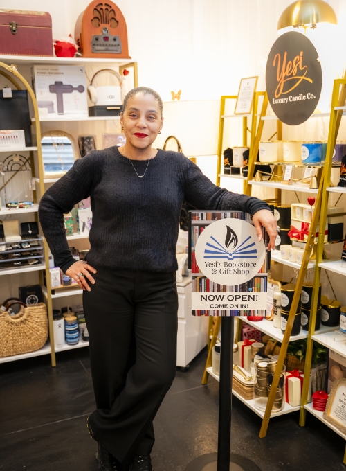 A woman wearing a black sweater and pants stands smiling in a shop. Shelves filled with various items, including baskets and candles, are in the background. She rests her hand on a sign that says Yesis Bookstore &amp; Gift Shop NOW OPEN! COME ON IN!