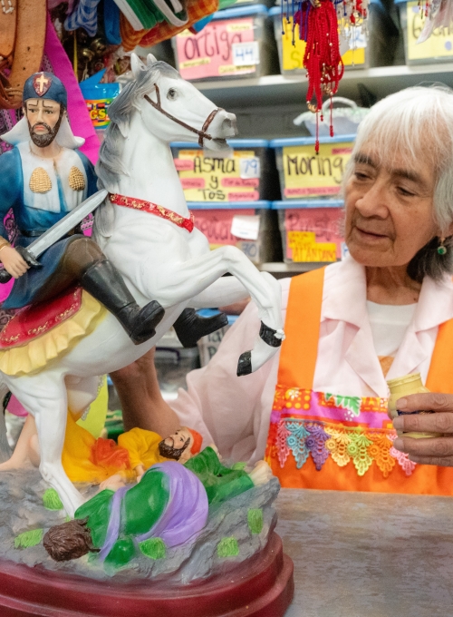 An elderly woman in an orange apron admires a colorful statue of a rider on a rearing white horse. The statue shows a man in medieval armor, with a figure in green lying on the ground. Shelves with labeled bins are in the background.