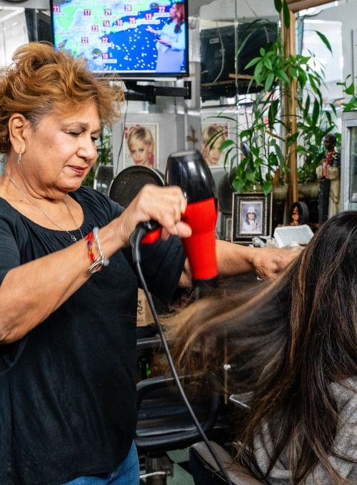 A woman blow-drying another persons hair in a salon. The stylist uses a red and black hairdryer. A TV in the background displays a weather map, and plants and framed photos are visible around the salon.