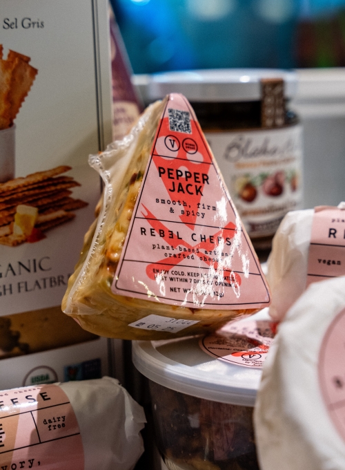 A wedge of Pepper Jack plant-based cheese with a pink label sits among various other food items. The label mentions smooth, firm & spicy and dairy free. Other labeled plant-based cheeses are partially visible around it. A wedge of Pepper Jack plant-based cheese with a pink label sits among various other food items. The label mentions smooth, firm & spicy and dairy free. Other labeled plant-based cheeses are partially visible around it.