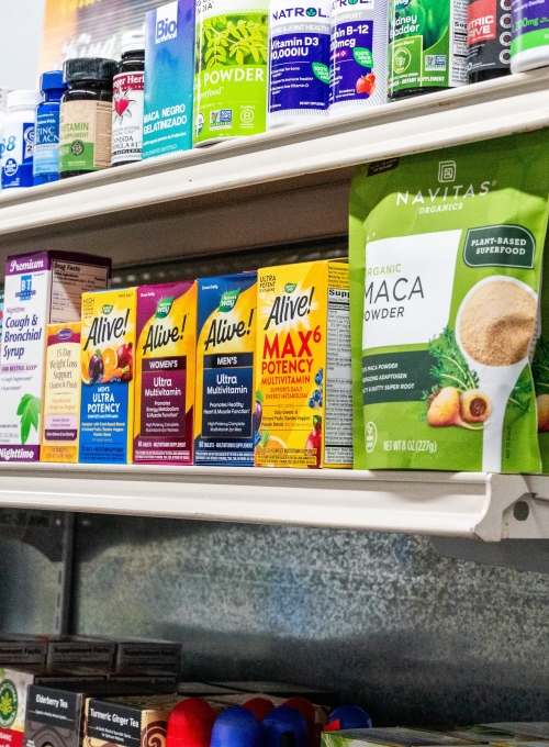 Shelves stocked with various vitamins, supplements, and health powders. Visible brands include Alive! multivitamins, maca powder, and Bio Synergy. The display features colorful packaging with different health product varieties.