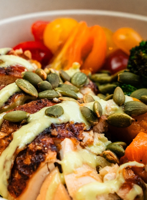 A close-up of a bowl with sliced roasted chicken topped with pumpkin seeds and drizzled with a light green sauce. Accompanied by vibrant cherry tomatoes and broccoli florets, creating a colorful and appetizing presentation. A close-up of a bowl with sliced roasted chicken topped with pumpkin seeds and drizzled with a light green sauce. Accompanied by vibrant cherry tomatoes and broccoli florets, creating a colorful and appetizing presentation.