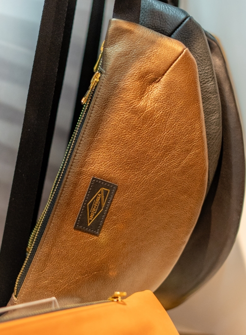 Close-up of a stylish sling bag with a metallic gold and black design. The bag features a diagonal zipper and a rectangular brand label. A soft focus hints at other bags in the background.