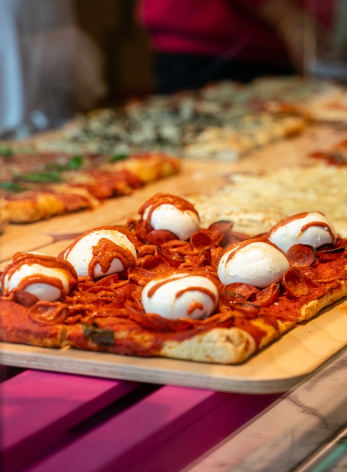 A slice of pizza topped with pepperoni, whole fresh mozzarella balls, and a drizzle of tomato sauce on a wooden board. Other pizzas are blurred in the background.