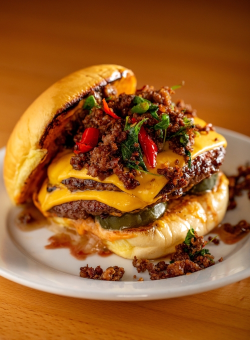 A cheeseburger with two patties, melted cheese, jalapeños, and a topping of crumbled sausage and herbs, served on a white plate. The bun is lightly toasted.