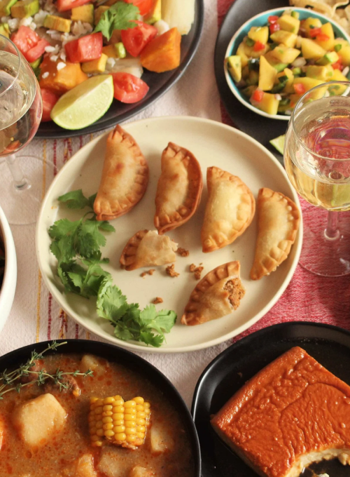 A table set with small empanadas on a white plate garnished with cilantro, surrounded by salad, soup with corn, flan dessert, and glasses of white wine.