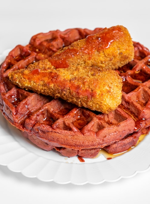 A plate with a red velvet waffle topped with two pieces of crispy fried chicken, drizzled with syrup, on a white scalloped-edge plate against a white background.