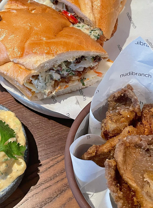 A sandwich filled with crispy chicken, greens, and sauce sits on paper labeled “nudibranch,” next to a bowl of fried chicken pieces and a dish of creamy dip garnished with parsley.