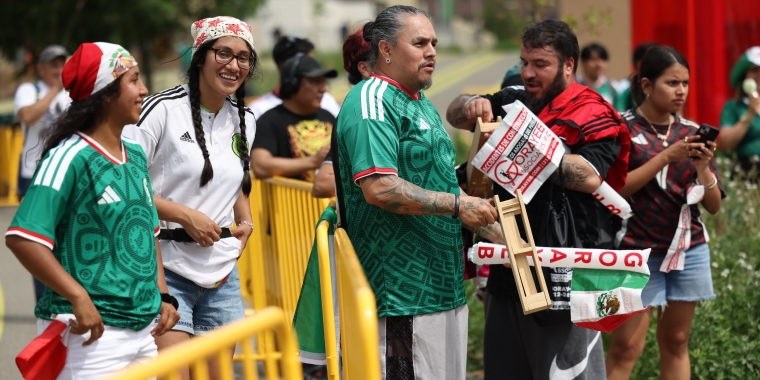 A group of soccer fans in green Mexico jerseys stand behind a yellow barrier, smiling and holding flags. One man holds a wooden noisemaker, and others wear festive hats and accessories. It is a festive outdoor setting.
