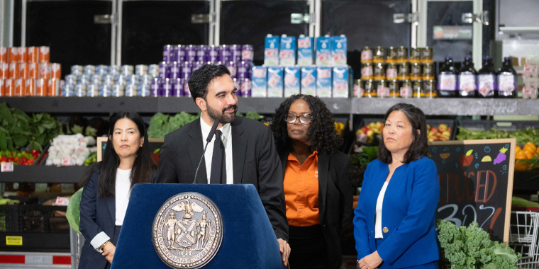 Mayor Mamdani speaks at a podium with the New York City seal in a grocery store with produce and products on shelves behind them.