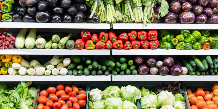 Shelves of fresh vegetables in a grocery store, including eggplants, leafy greens, peppers, cucumbers, tomatoes, and cabbages, all neatly arranged in rows.