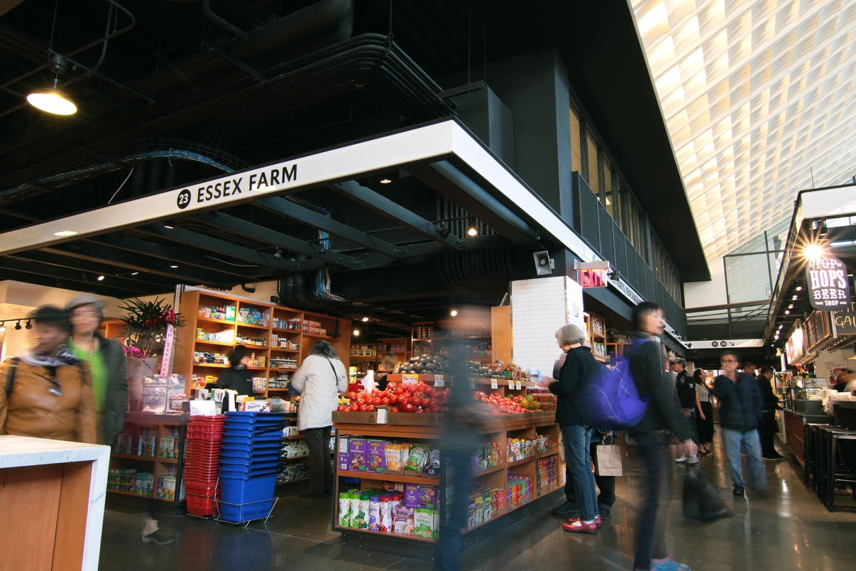 A bustling Essex Market features a variety of colorful fruit and packaged goods. Shoppers move about under signage reading Essex Farm. The atmosphere is lively and vibrant, with people browsing and walking around. Photo by Nina Loschiavo.