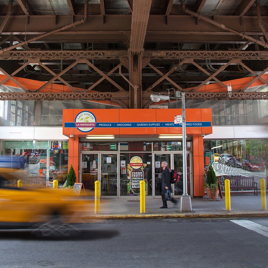 A bright orange storefront with a sign reading La Marqueta sits under a steel bridge. A blurred yellow taxi passes in the foreground, while a person in black walks by the entrance, which displays various signs and advertisements.