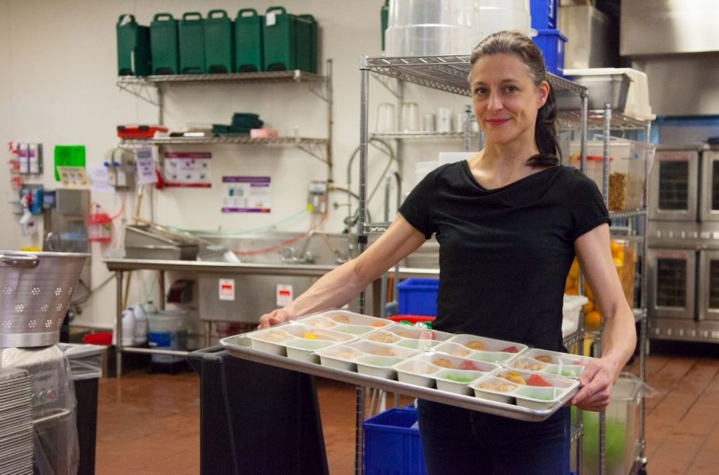 A person in kitchen of Little Green Gourmets at La Marqueta Market holds a tray of colorful, portioned meals. Shelves filled with kitchen supplies and ingredients are visible in the background. The setting is clean and organized.