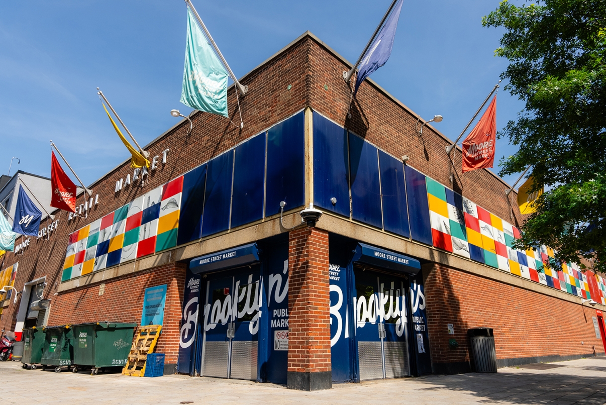 Outdoor view of Moore Street Market building with colorful flags and signs. The building has brick walls and large windows. 