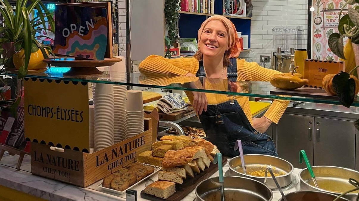 A smiling person wearing a yellow shirt and overalls stands behind a counter in a cozy café. The counter displays pastries and soup in pots. Bright decorations and colorful books adorn the background.