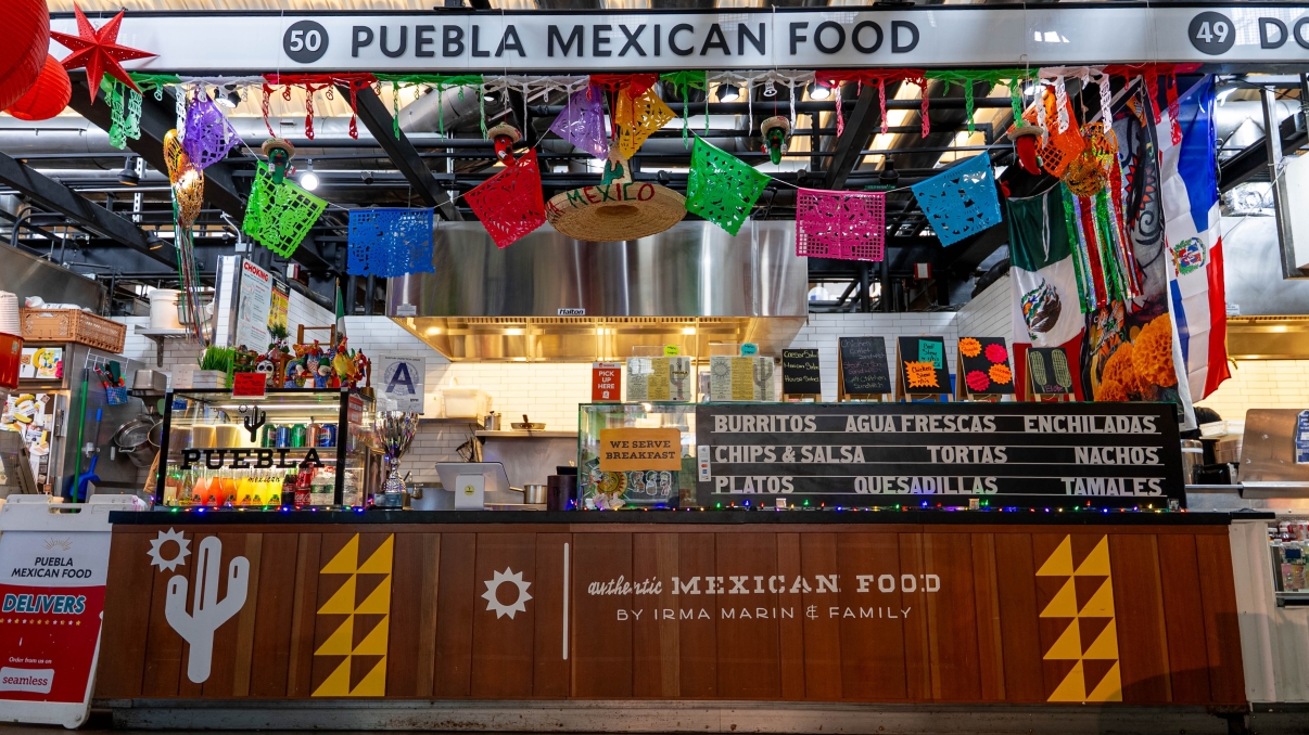 A colorful Mexican food stall with vibrant papel picado banners, a counter displaying a menu featuring burritos, tacos, enchiladas, and tamales. Decor includes piñatas and festive lights, with signs in English and Spanish.