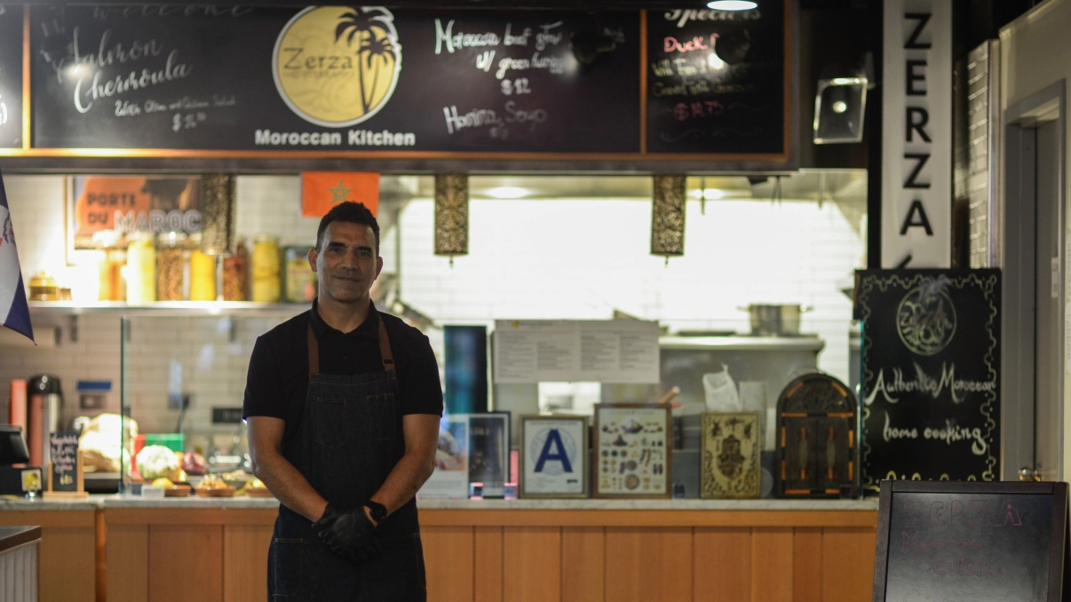 Man in a black shirt and apron standing in front of a Moroccan food counter named Zerza. The counter displays menus and a Moroccan flag, with warm lighting and various decorative elements.