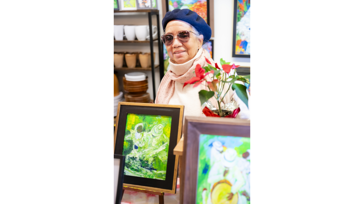Elderly person wearing a blue beret and sunglasses, standing in front of a table with framed colorful paintings. They are dressed in a pink scarf and white sweater, with a red poinsettia plant beside them. Shelves with dishes are in the background.