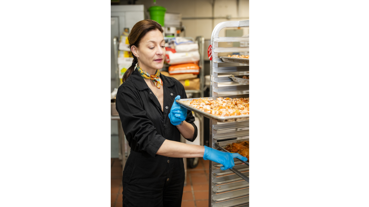 A woman in a black outfit and blue gloves is placing a baking tray with food onto a rack in a commercial kitchen. She appears focused on her task. Various kitchen supplies and equipment are visible in the background.