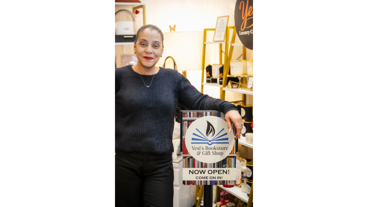A woman smiling and standing in a bookstore, holding a sign that reads Yesis Bookstore &amp;amp; Gift Shop, NOW OPEN! COME ON IN! The store has shelves with books and gift items in the background.